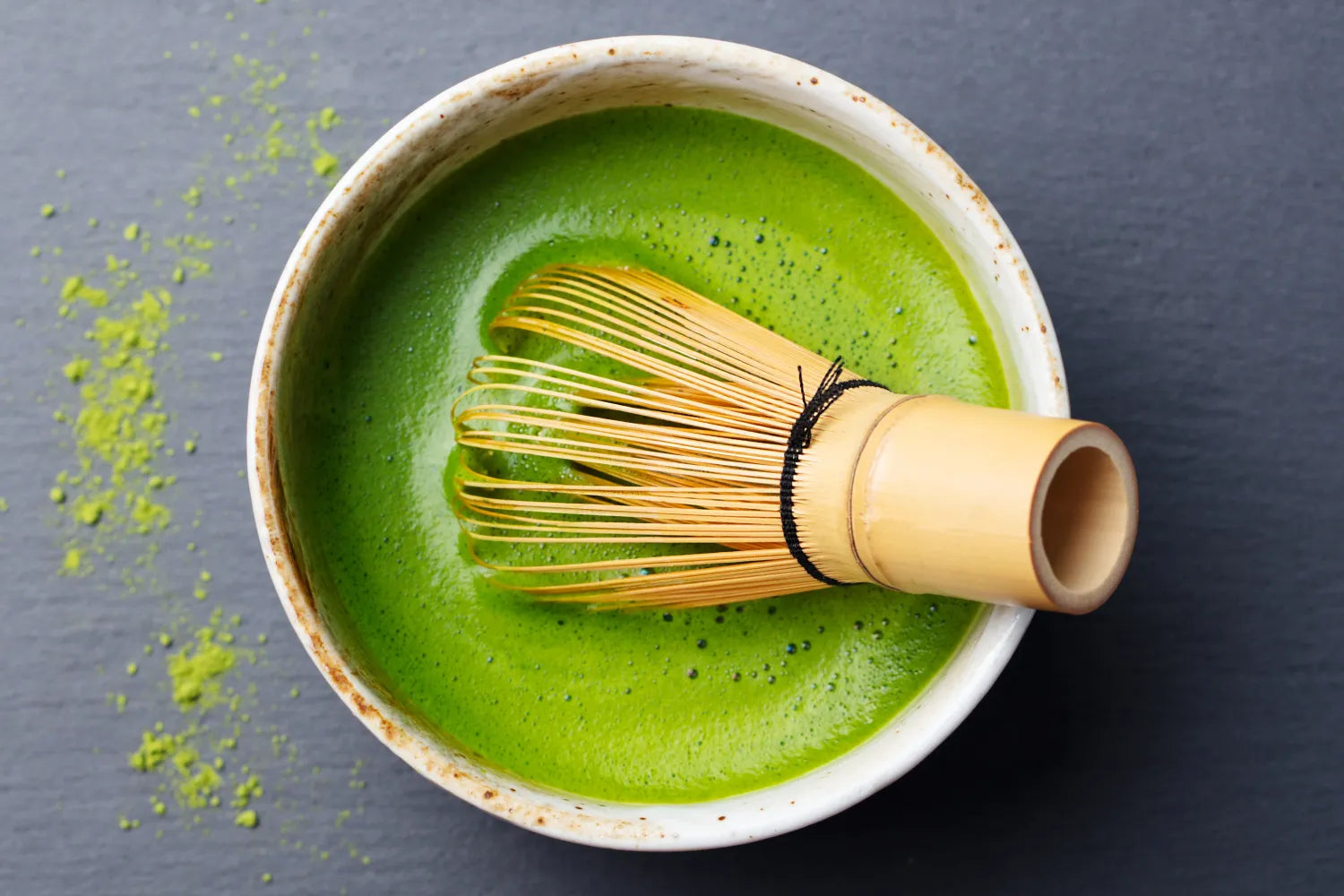 Top-down view of a bowl of bright green ceremonial-grade matcha being whisked with a bamboo chasen on a dark slate surface