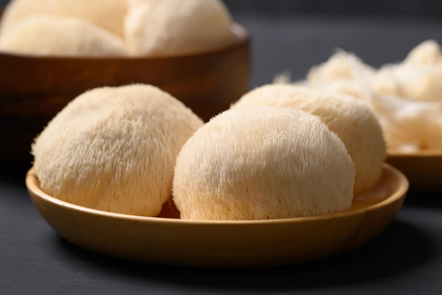 Close-up of three Lion’s Mane mushrooms showing their white shaggy spines on a wooden plate against a dark background