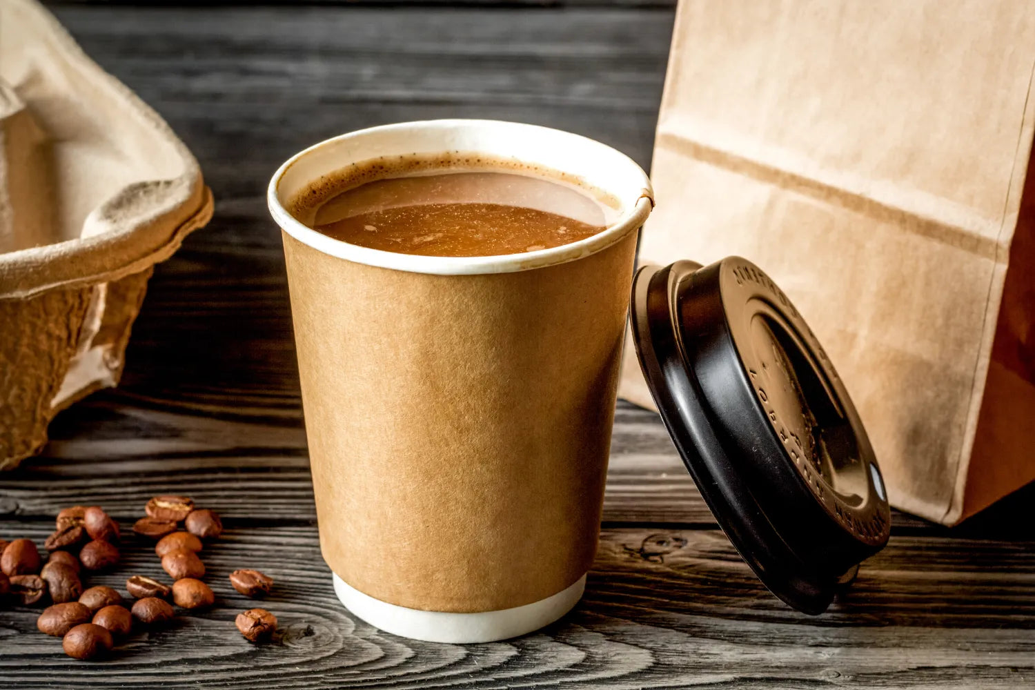 Takeaway coffee in a brown paper cup on a wooden table with scattered coffee beans, a black lid leaning against a paper bag