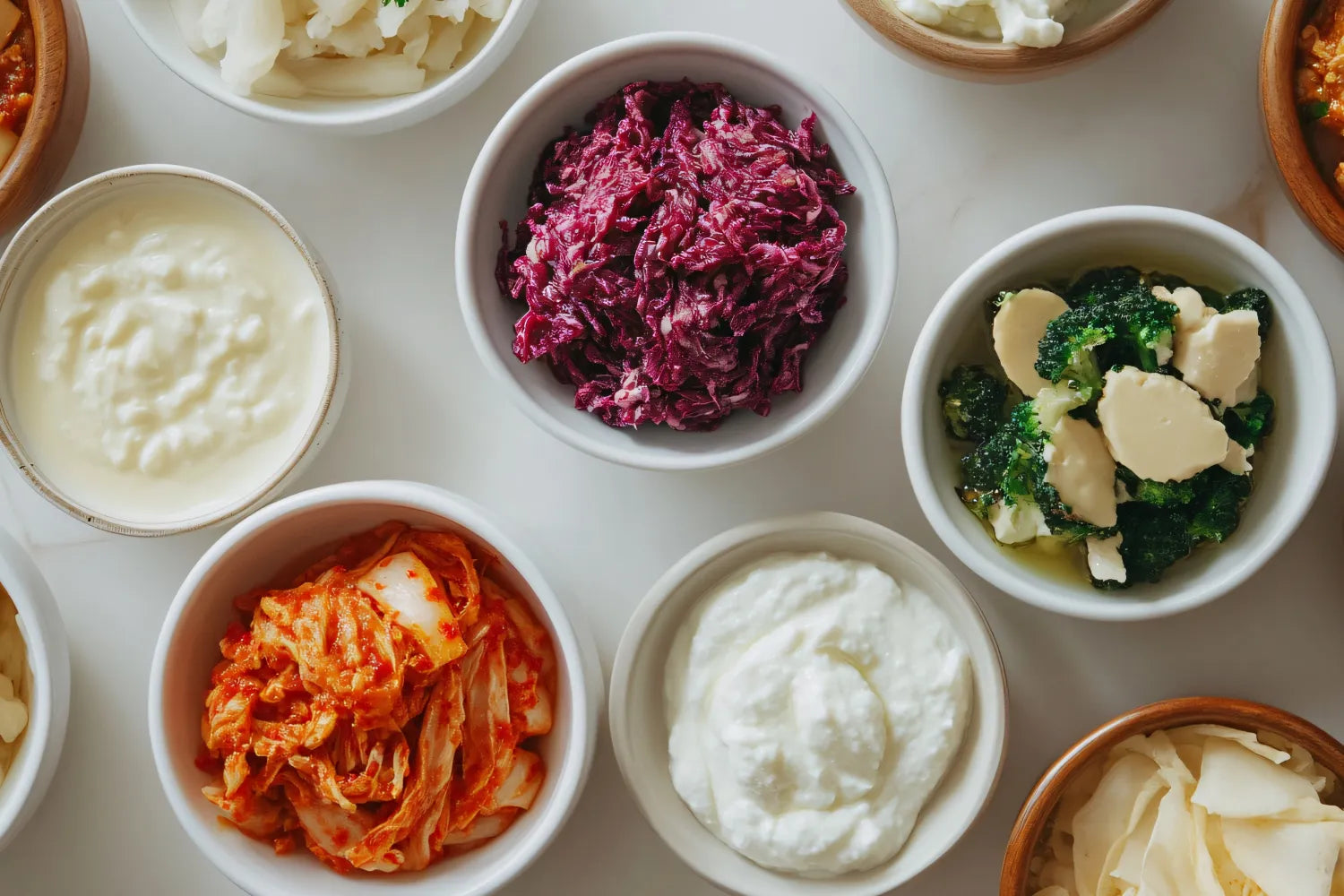 Bowls of gut-friendly fermented foods arranged on a white surface, including kimchi, red cabbage sauerkraut, yogurt, and seasoned greens