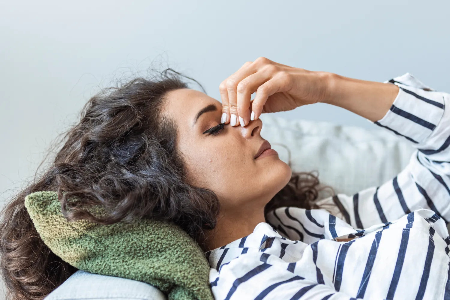 Woman lying on a couch pinching the bridge of her nose as if experiencing a headache, striped shirt and green pillow behind her