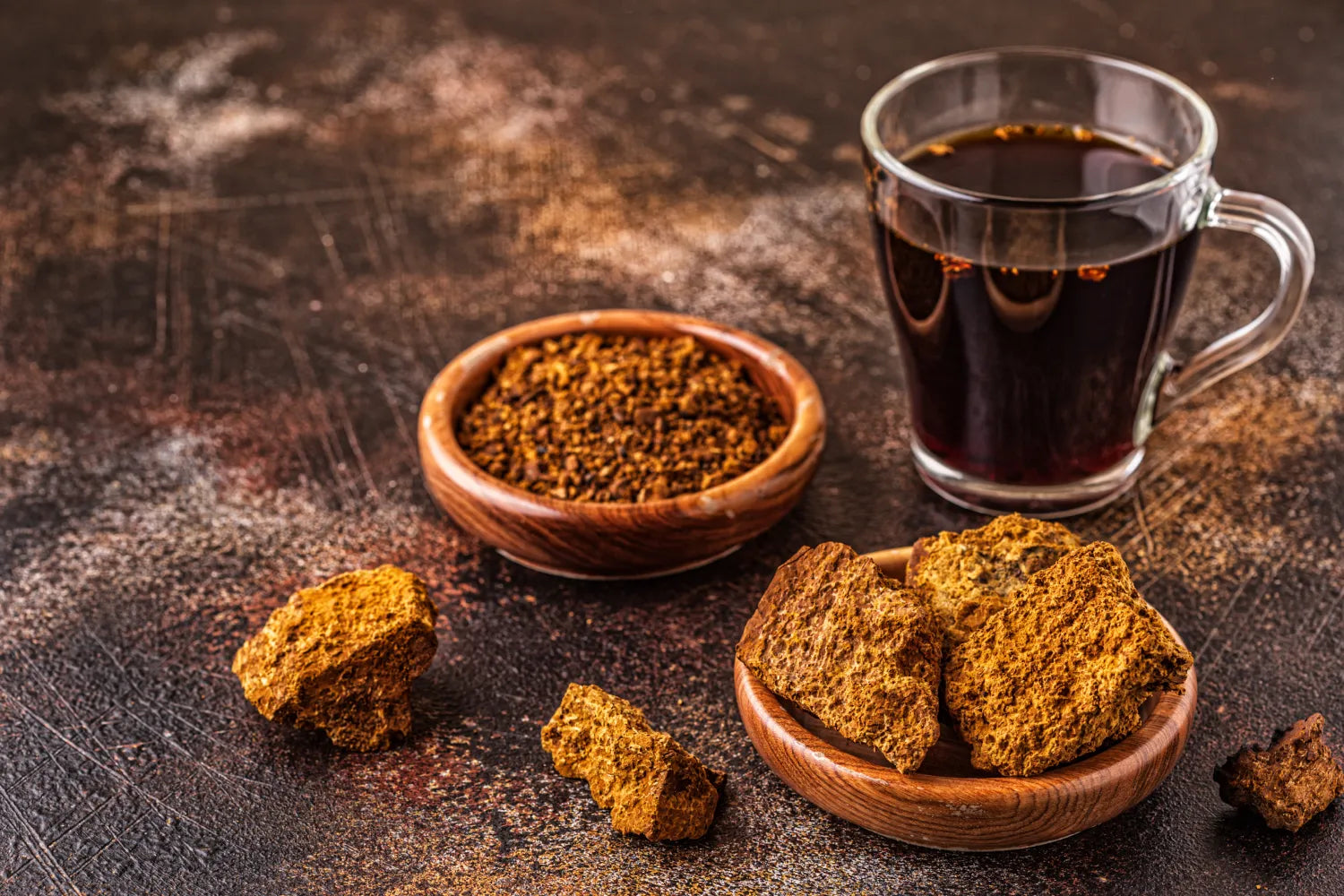 Glass mug of black coffee beside wooden bowls of chaga chunks and ground chaga on a dark textured surface