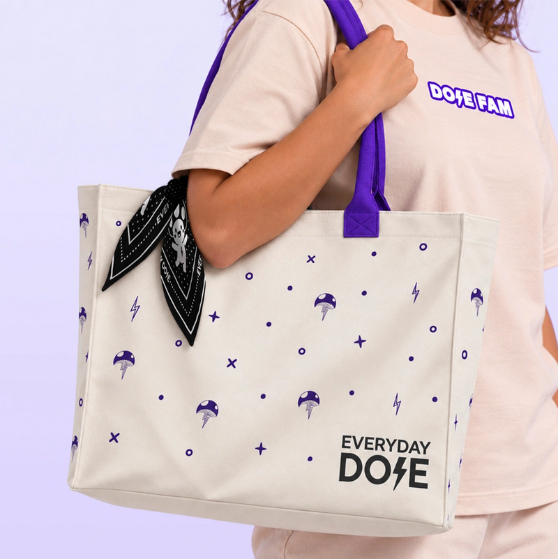 Cream tote bag with purple handles and mushroom and lightning bolt print carried by a woman with a black patterned bandana attached