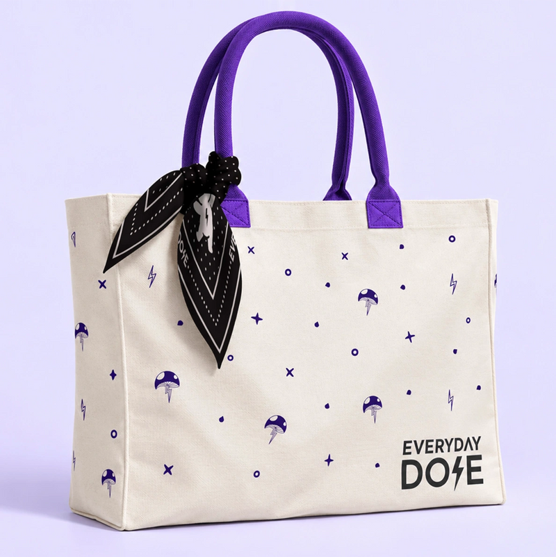 Cream canvas tote bag with purple handles and a black patterned scarf tied to one side against a lavender background