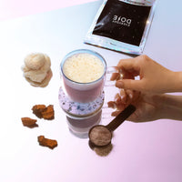 Hands holding a glass cup of frothy beverage beside a spoonful of brown powder and scattered brown pieces on a pastel surface