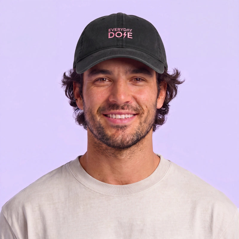 Smiling man wearing a black baseball cap with pink embroidered lettering and a light beige shirt against a lavender background