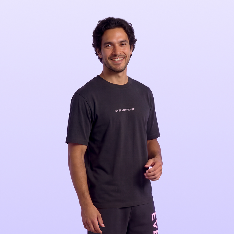 Smiling man wearing a black Everyday Dose T shirt and matching pants with pink vertical lettering against a soft lavender background