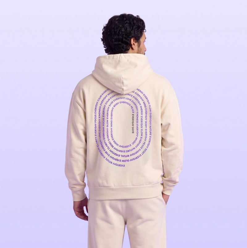 Model wearing a cream hooded sweatshirt with purple circular back lettering and matching pants against a soft lavender background