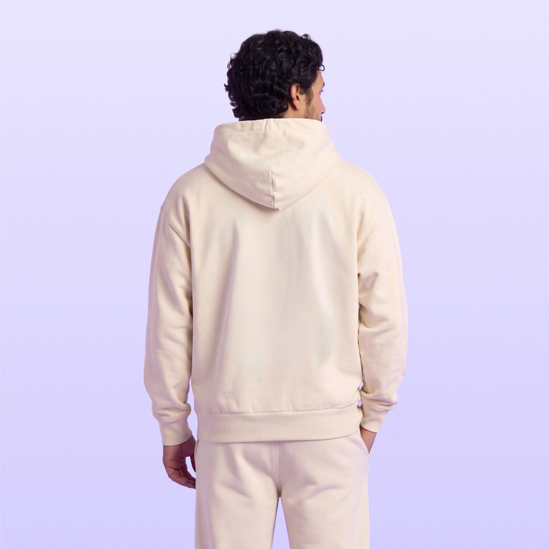 Back view of a man wearing a cream hooded sweatshirt and matching pants against a soft lavender background