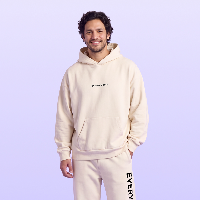Smiling man wearing a cream hoodie and matching sweatpants with Everyday Dose branding against a soft lavender background