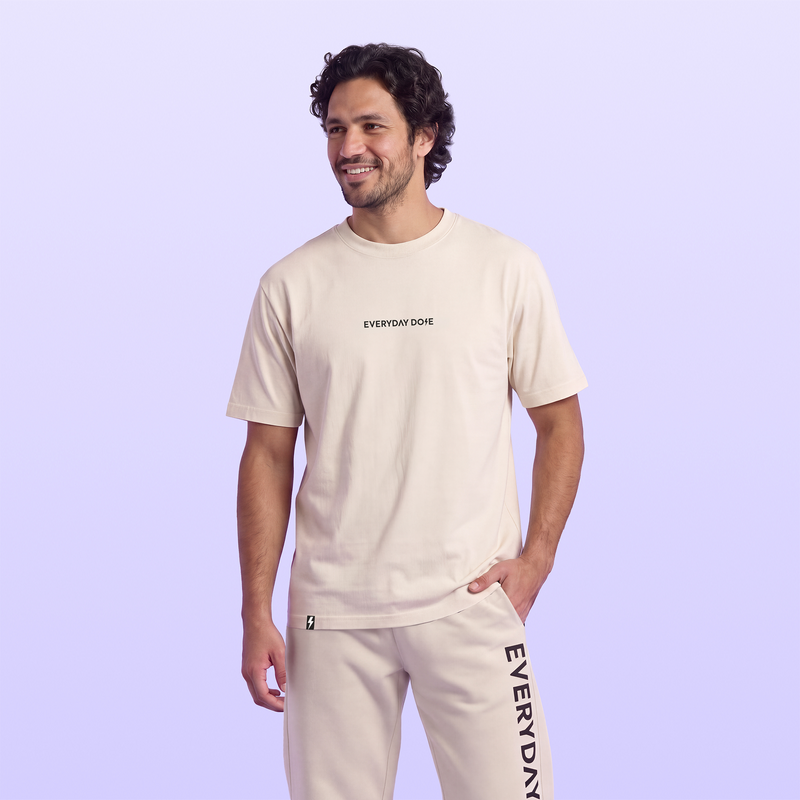 Smiling man wearing a cream Everyday Dose T shirt and matching sweatpants with vertical black lettering against a lavender background