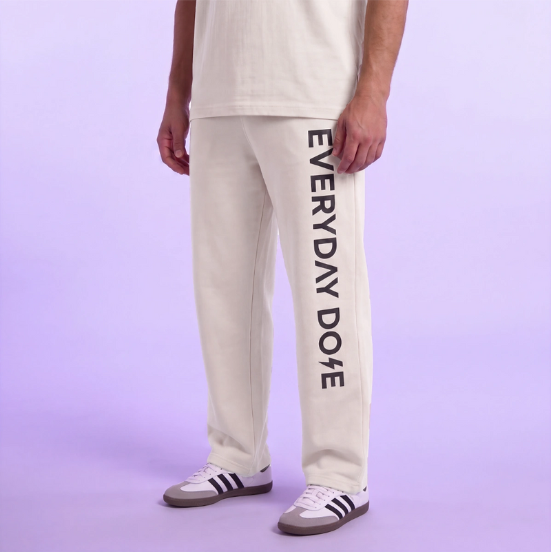 Model wearing cream sweatpants with vertical black Everyday Dose lettering and a matching shirt with striped sneakers against a soft lavender background
