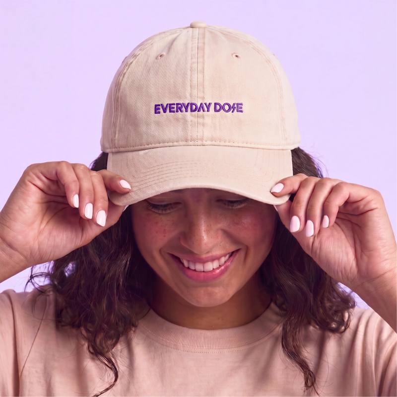 Smiling woman adjusting a beige Everyday Dose baseball cap against a soft lavender background