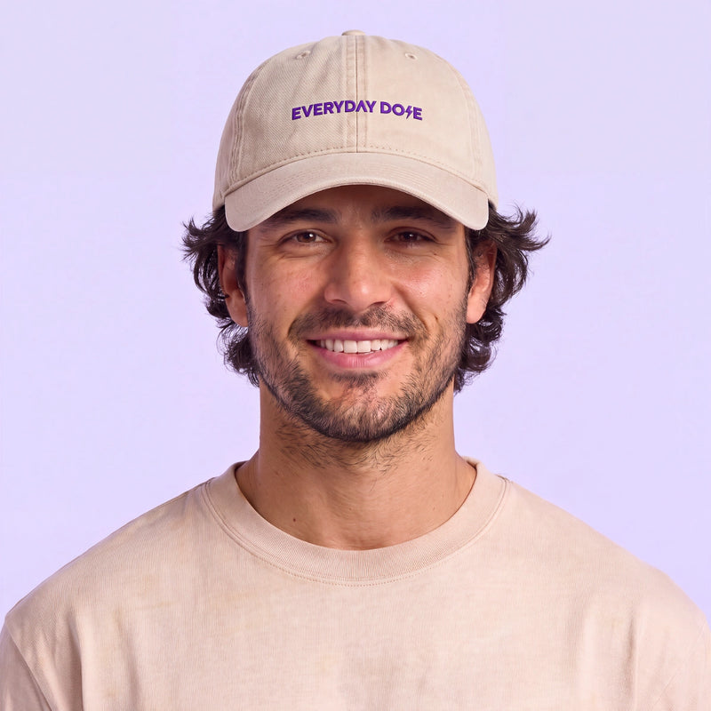 Smiling man wearing a beige baseball cap with purple Everyday Dose lettering against a soft lavender background