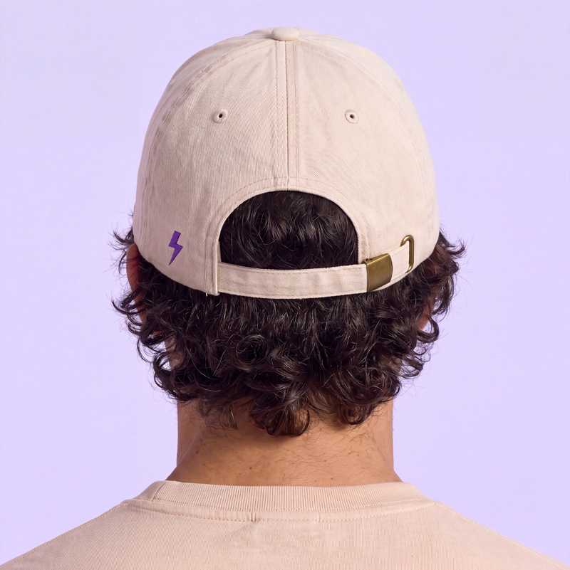 Back view of a person wearing a beige adjustable cap with a purple lightning logo against a soft lavender background