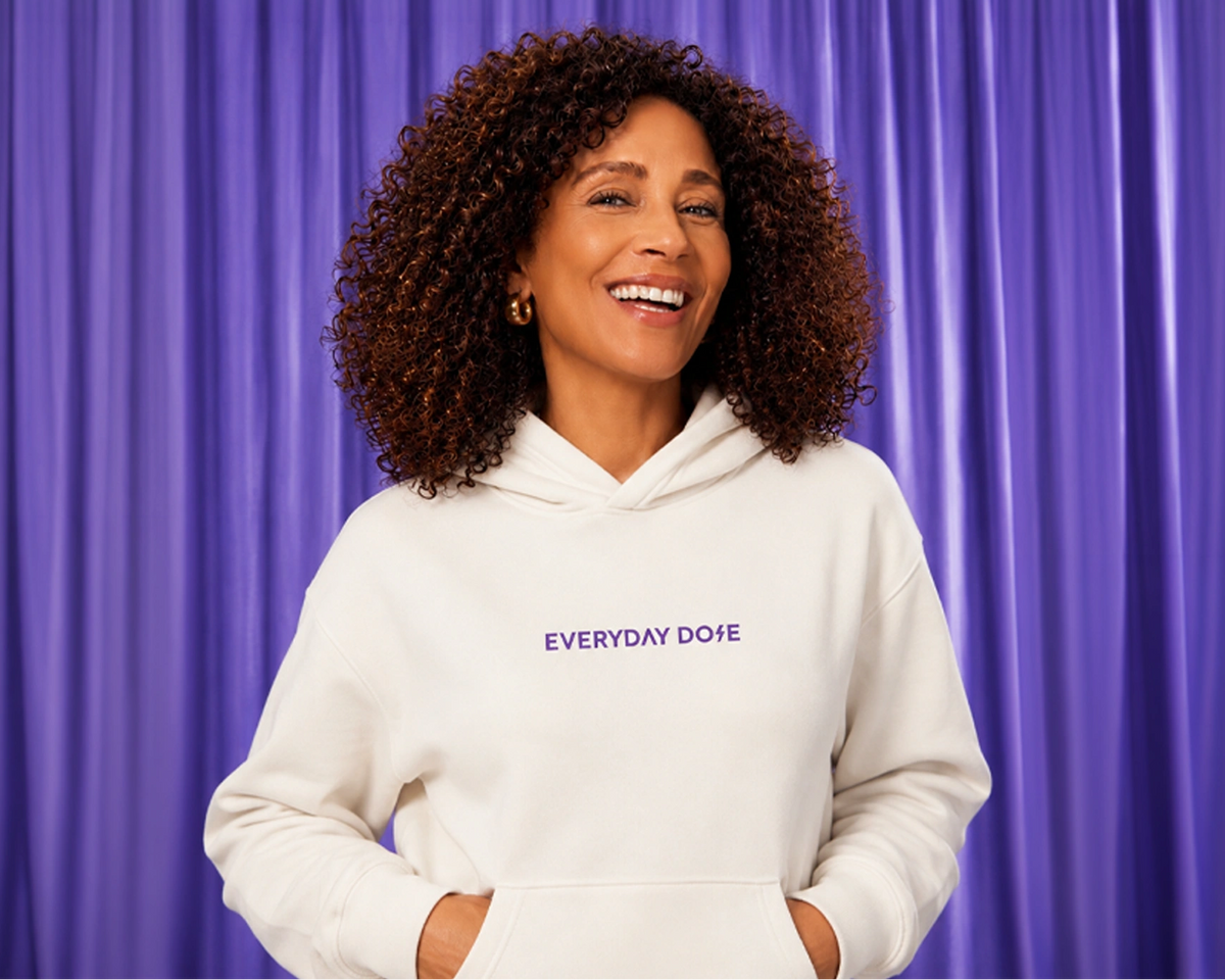 A smiling woman with curly brown hair wearing a white hoodie with the text 'EVERYDAY DOSE' in purple on the chest, standing in front of a purple curtain backdrop.