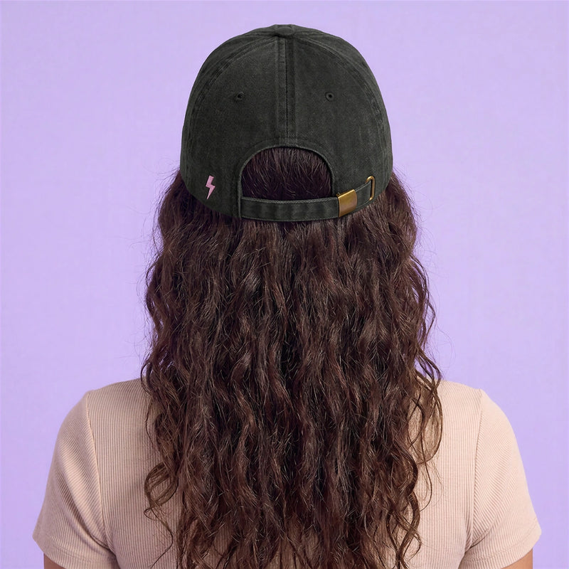Back view of a model wearing a black adjustable cap with a small lightning detail against a soft lavender background