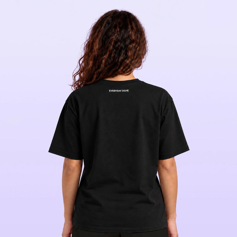 Woman seen from behind wearing a black short sleeve shirt with tiny Everyday Dope lettering near the collar against a light lavender backdrop