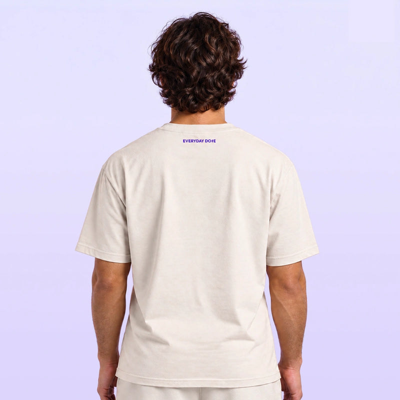 Man viewed from behind wearing a cream short sleeve T shirt with tiny purple Everyday Dope lettering below the collar on a lavender backdrop