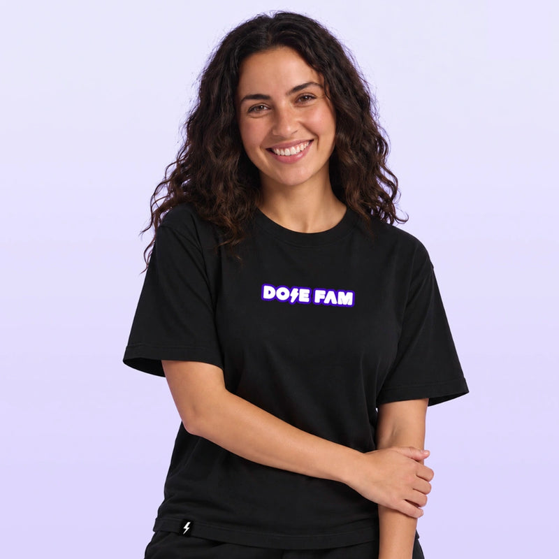 Smiling woman modeling a black T shirt with a colorful DOSE FAM logo against a soft lavender background