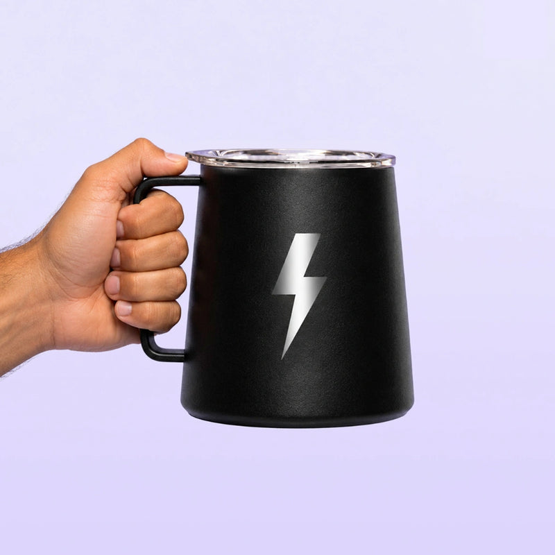 Black insulated travel mug with a handle and silver lightning bolt emblem held by a hand against a soft lavender background