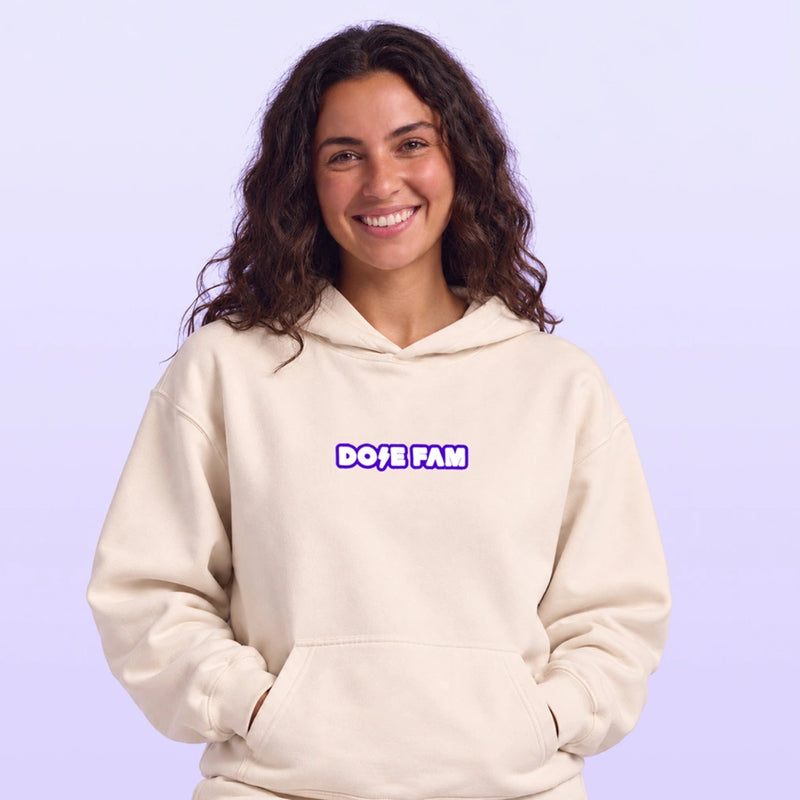 Smiling woman wearing a cream pullover hoodie with a purple and white chest logo against a soft lavender background