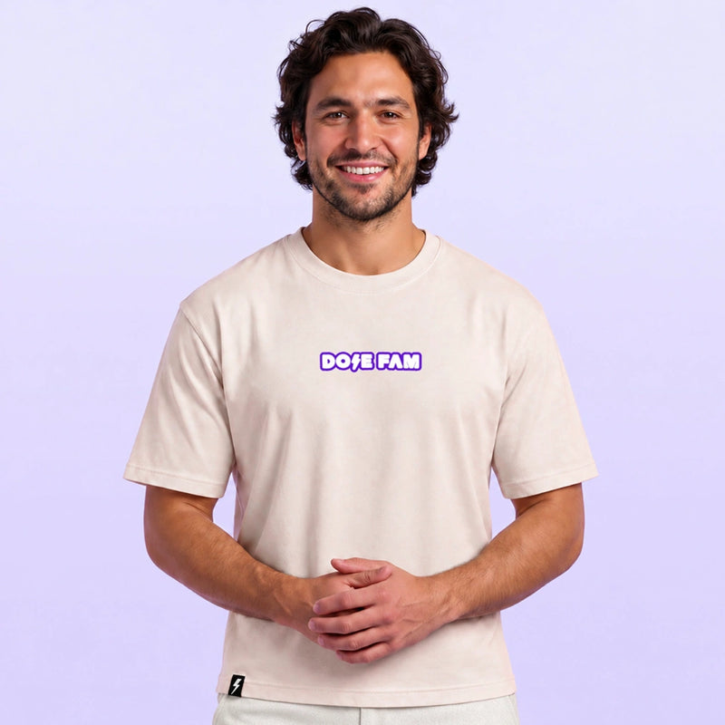 Smiling man modeling a cream short sleeve T shirt with a purple and white DOSE FAM logo against a lavender background