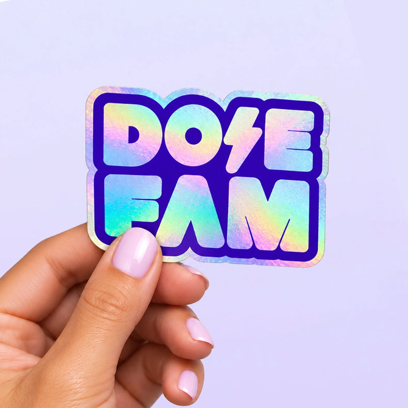 Holographic Dose Fam sticker with bold blue lettering held by a hand with lavender painted nails against a pale lavender background