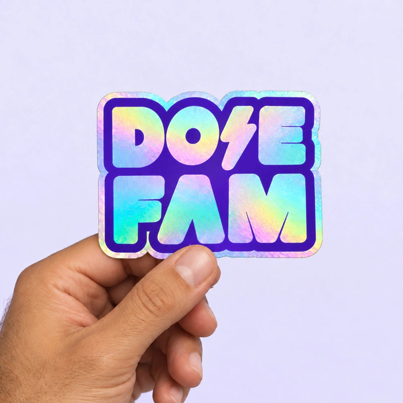 Holographic DOSE FAM sticker held between fingers against a soft lavender background