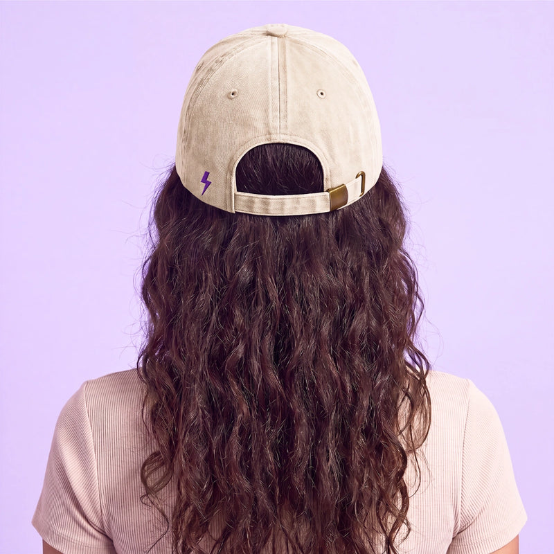 Back view of a person with long wavy hair wearing a beige baseball cap with a purple lightning bolt detail against a lavender background