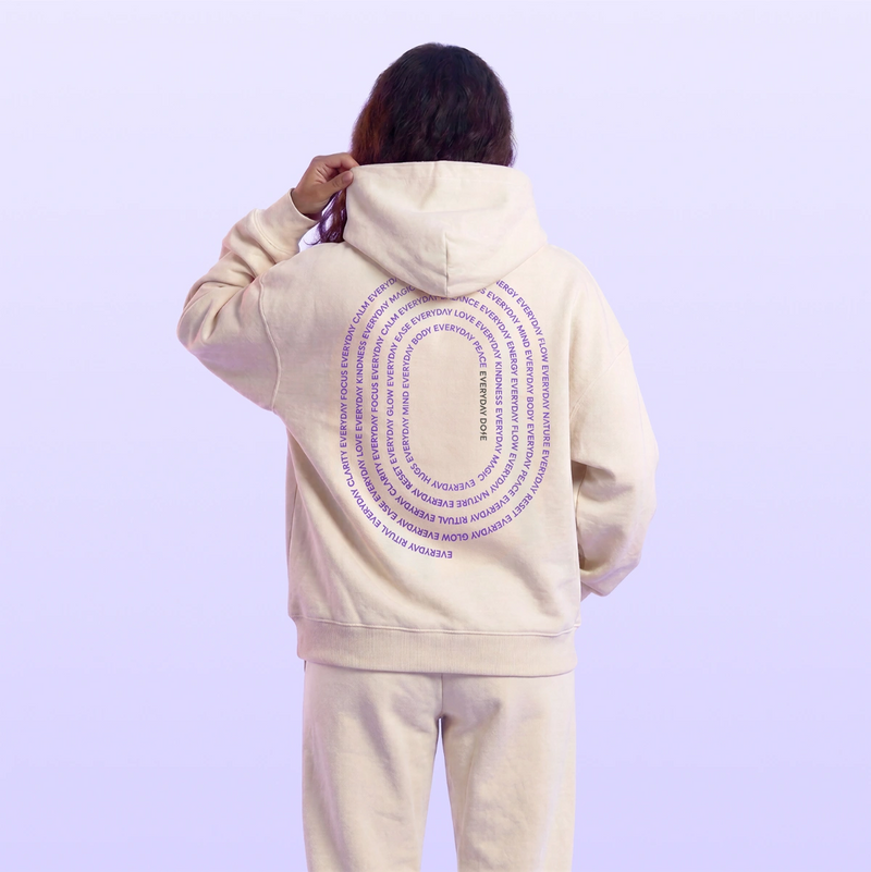 Back view of a model wearing a cream hooded sweatshirt and matching pants with purple circular text artwork against a pale lavender background