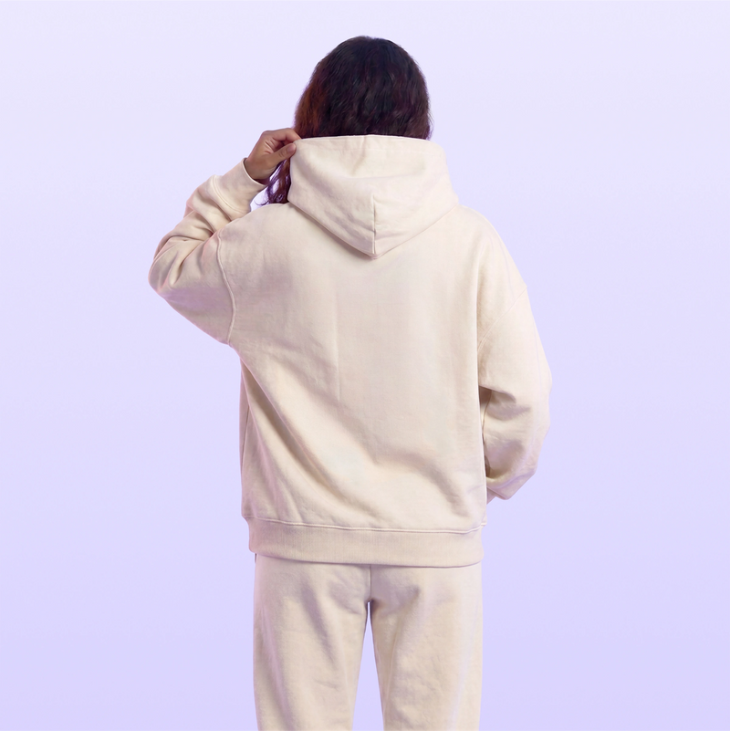 Back view of a model wearing a cream hooded sweatshirt and matching sweatpants against a soft lavender background
