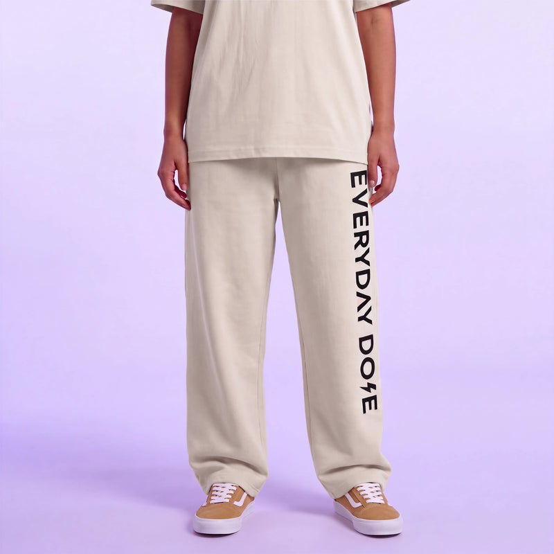 Model wearing a cream T shirt and matching sweatpants with vertical black Everyday Dose lettering and brown sneakers against lavender background