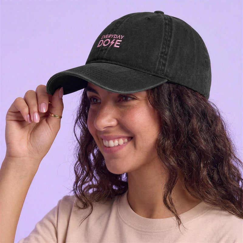 Smiling woman adjusting a black cap with an embroidered Everyday Dose logo while wearing a light pink shirt against a lavender background