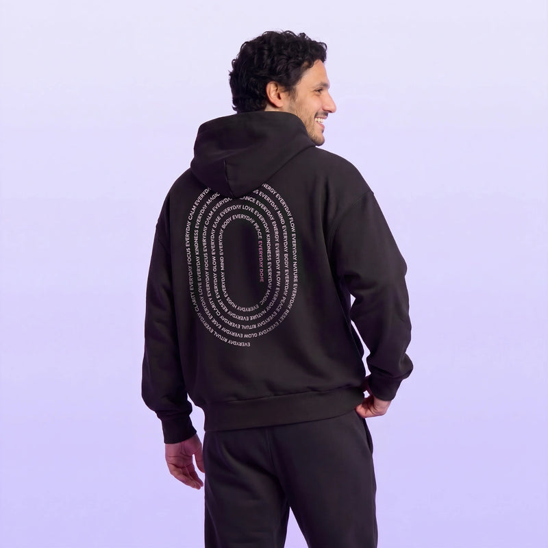Man wearing a black hooded sweatshirt with white oval text artwork and matching pants against a soft lavender background