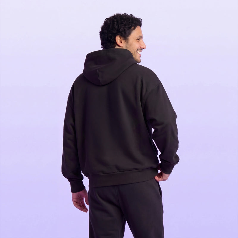 Back view of a smiling man wearing a black hooded sweatshirt and matching joggers against a soft lavender background