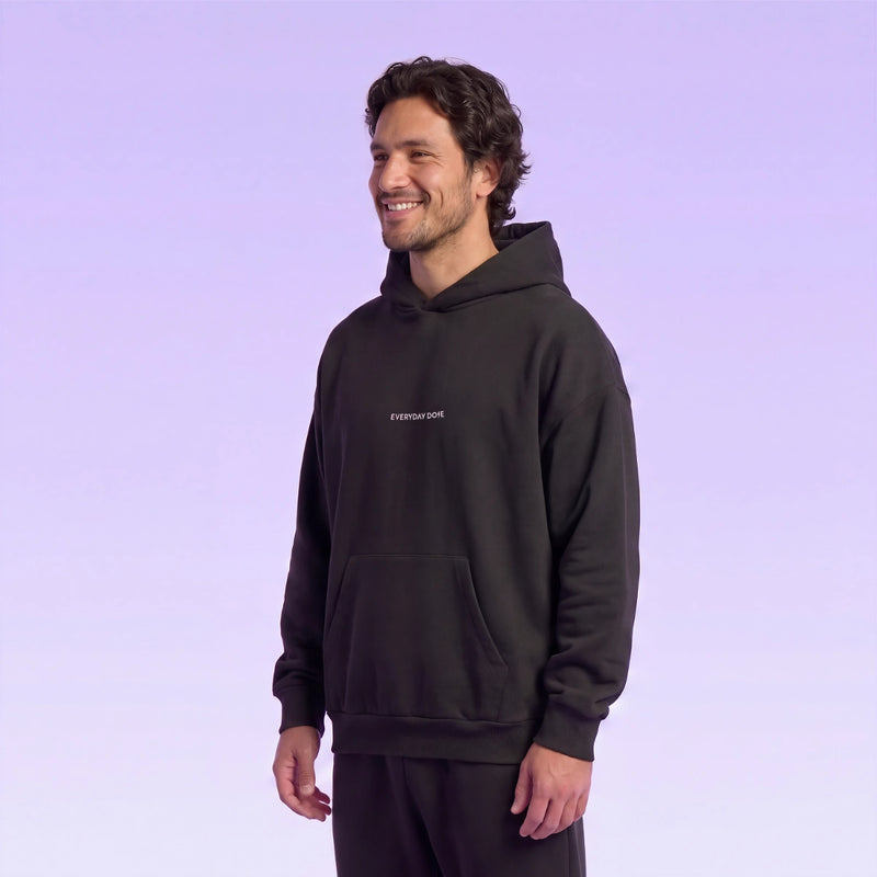 Smiling model wearing a black hooded sweatshirt with a small chest logo against a soft lavender background