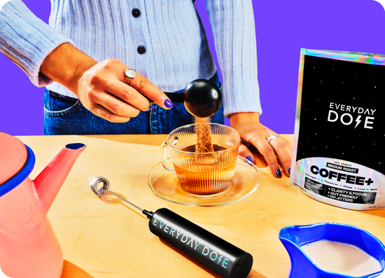 Hands scooping Everyday Dose Coffee Plus into a glass mug with branded USB frother and pouch on a wooden table