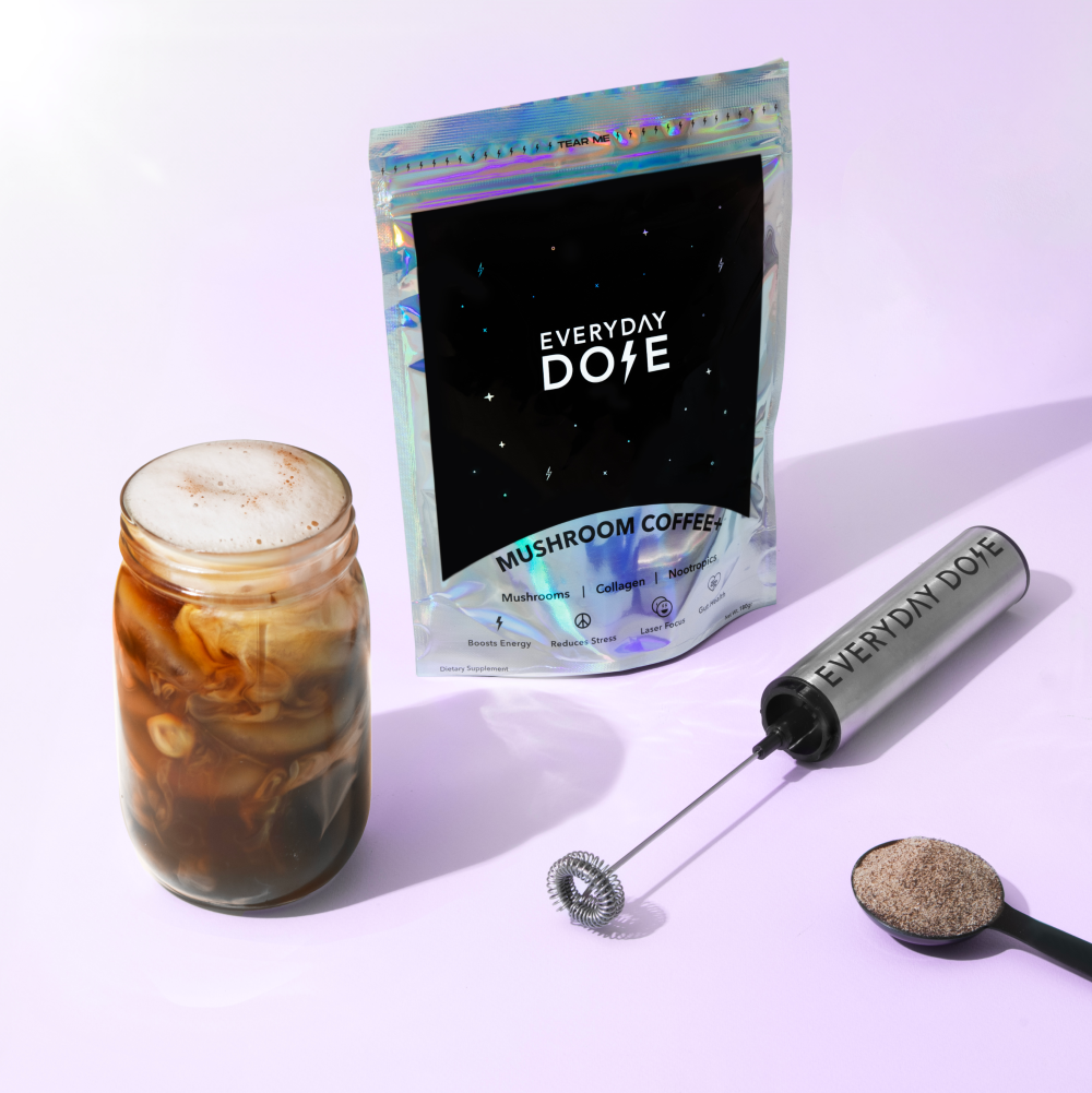 Photo of the Everyday Dose mushroom coffee+ 30 serving bag, the rechargeable frother, gunmetal serving scooper, and a mason jar with mushroom coffee+ in it on a purple background.